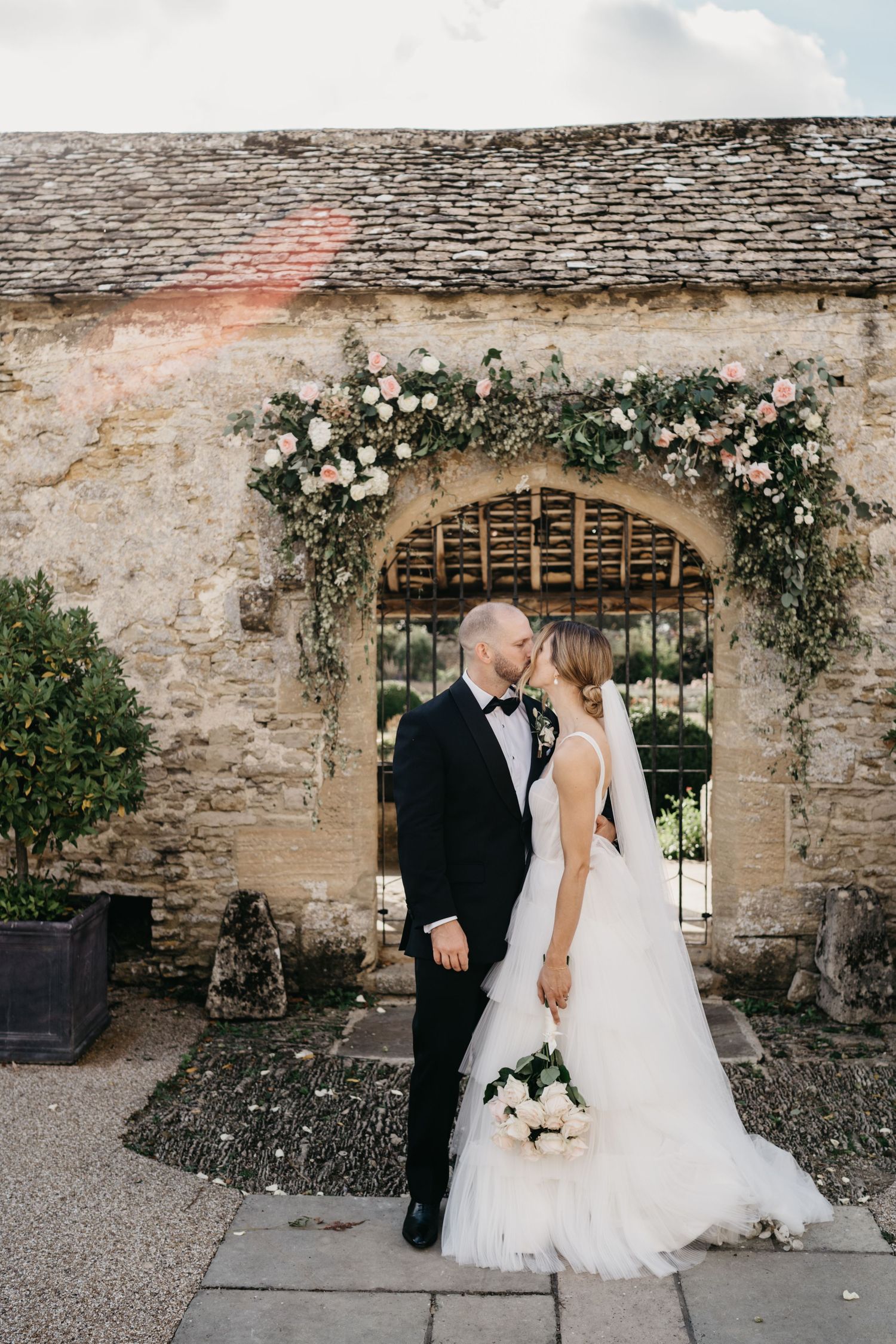 A romantic wedding scene at a rustic stone wall with climbing white roses and an arched doorway.