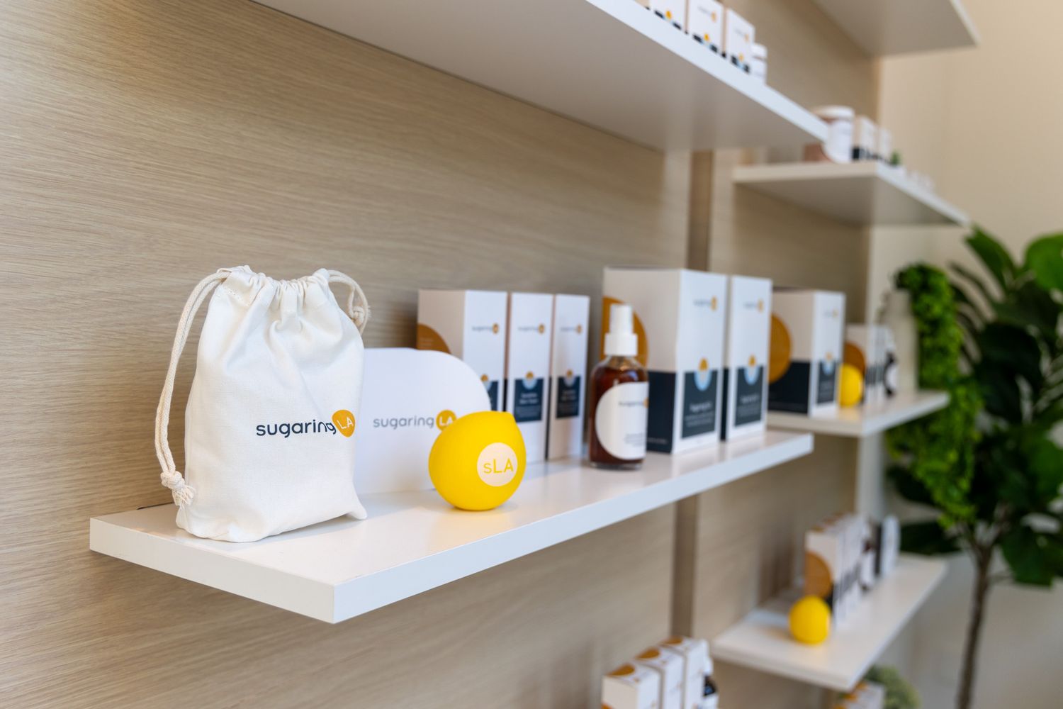 White product bottles and packages displayed on modern wooden shelves with yellow accent decorations and green plants. sugaringLA alternativ