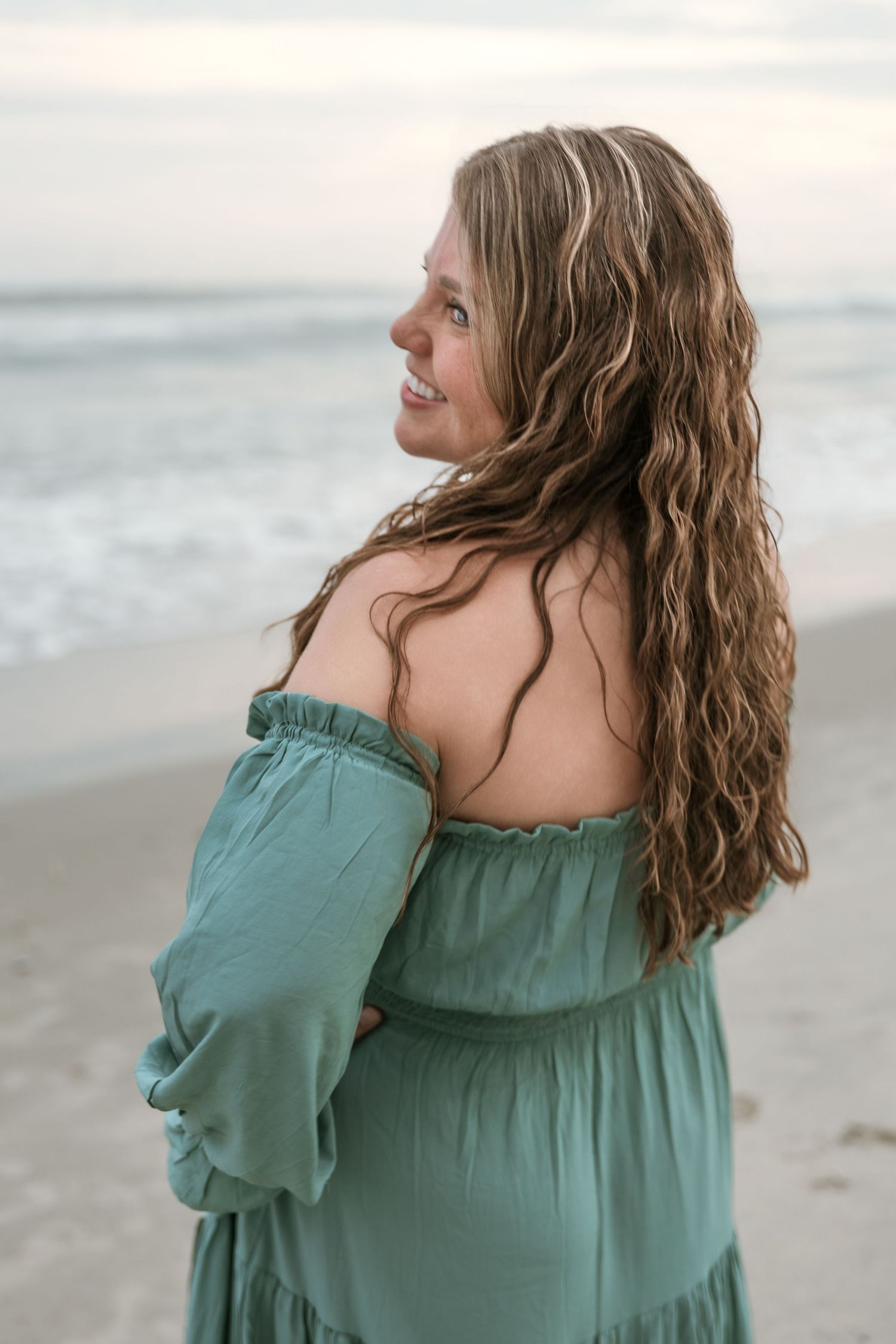 A woman in a teal off-shoulder dress gazes thoughtfully at the ocean during a beach photoshoot.