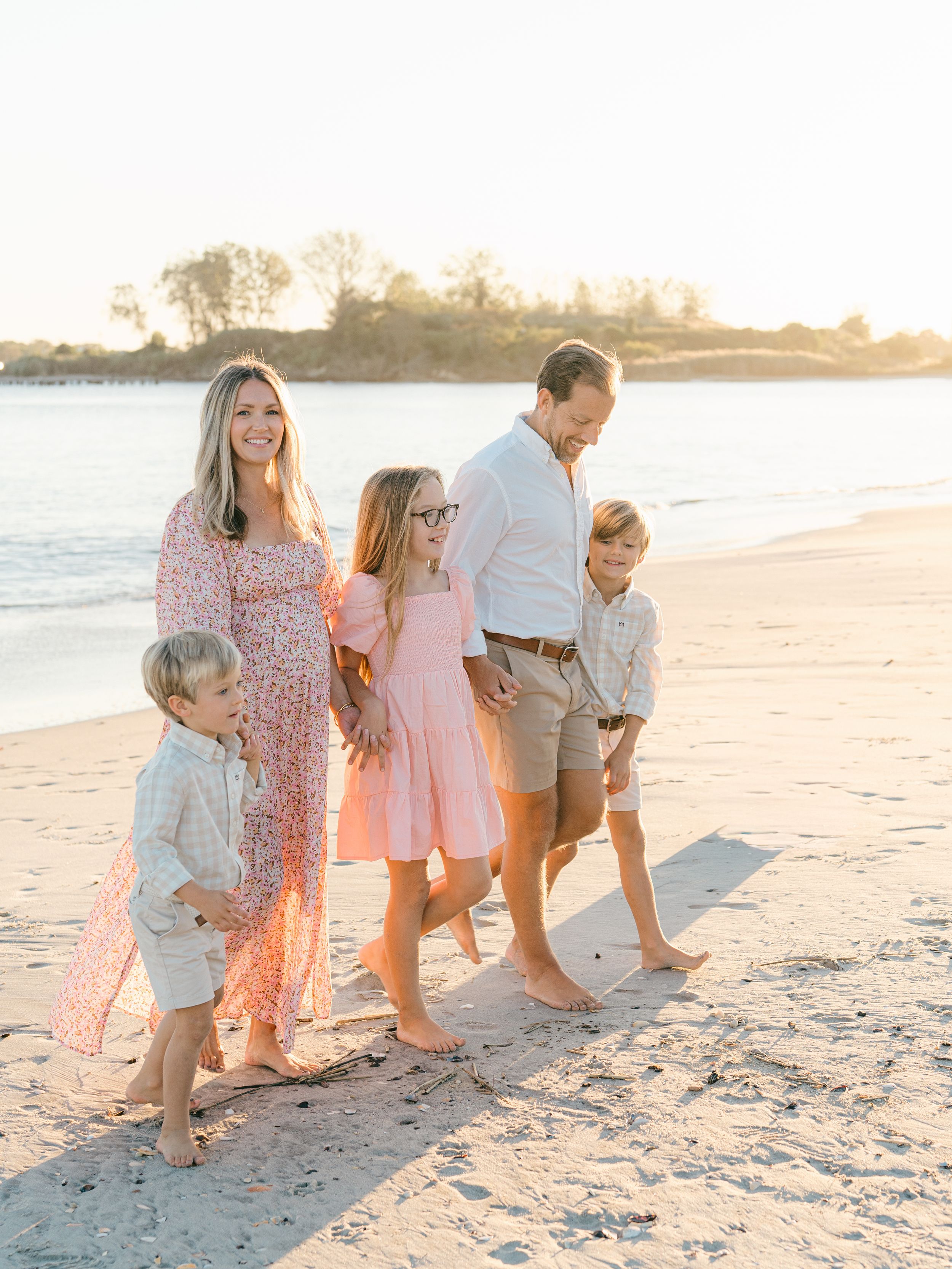 a family dressed in pinks and tans walking down the manasquan beach with the sun setting in the background.