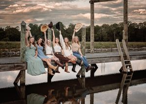 Wedding party members sit casually on a wooden dock at sunset creating a relaxed group photo atmosphere.