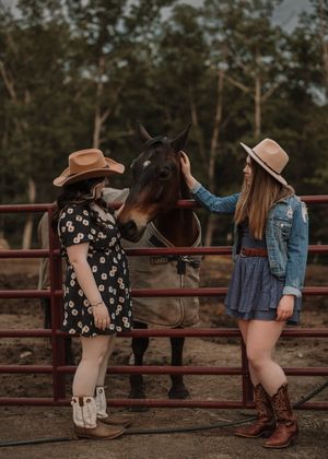 Two friends in cowboy boots sit casually on a red fence while visiting with a dark horse at a ranch.