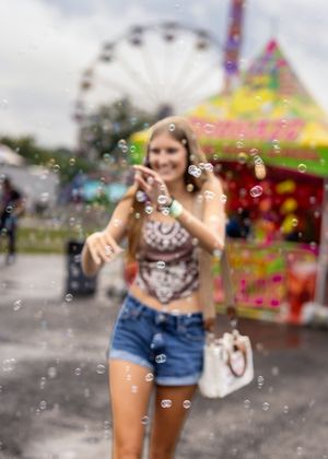 Soap bubbles float in front of a carnival scene with a Ferris wheel and colorful attractions in the background.