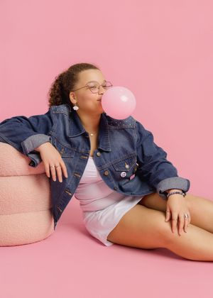 Someone wearing denim jacket and white top blows pink bubblegum while lounging against pink background.