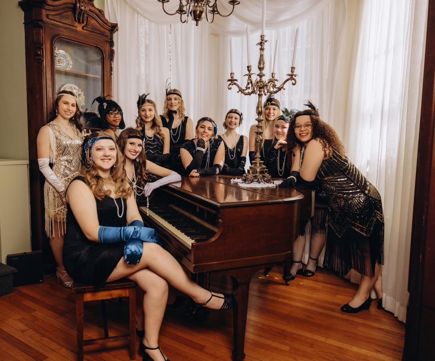 Group of people in formal black attire posing together in an elegant room with hardwood floors and white curtains.