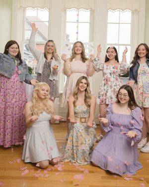 A group in floral and pastel dresses celebrates joyfully in a bright sunlit room with scattered rose petals on the floor.