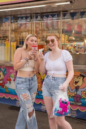 Two people in denim shorts stand together at a carnival with colorful artwork visible in the background.