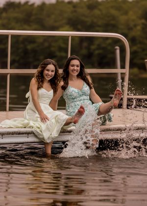 Two friends in dresses splash their feet in a lake during a playful summer photoshoot at sunset.
