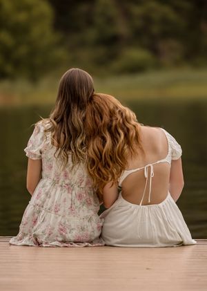 Two women in white and floral dresses sitting side by side on a wooden dock by a tranquil lake at sunset.
