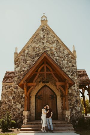 Couple embracing before a rustic stone chapel with wooden arched doorway and cross on top.