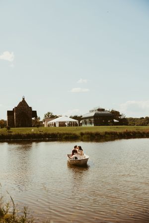 Couple sharing a romantic moment in a rowboat on a serene pond with a wedding venue and historic chapel in the background.
