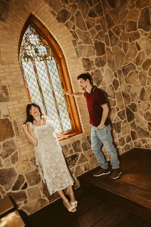 Couple posing by a Gothic stained glass window in a stone chapel interior, woman in white dress, man in red polo.