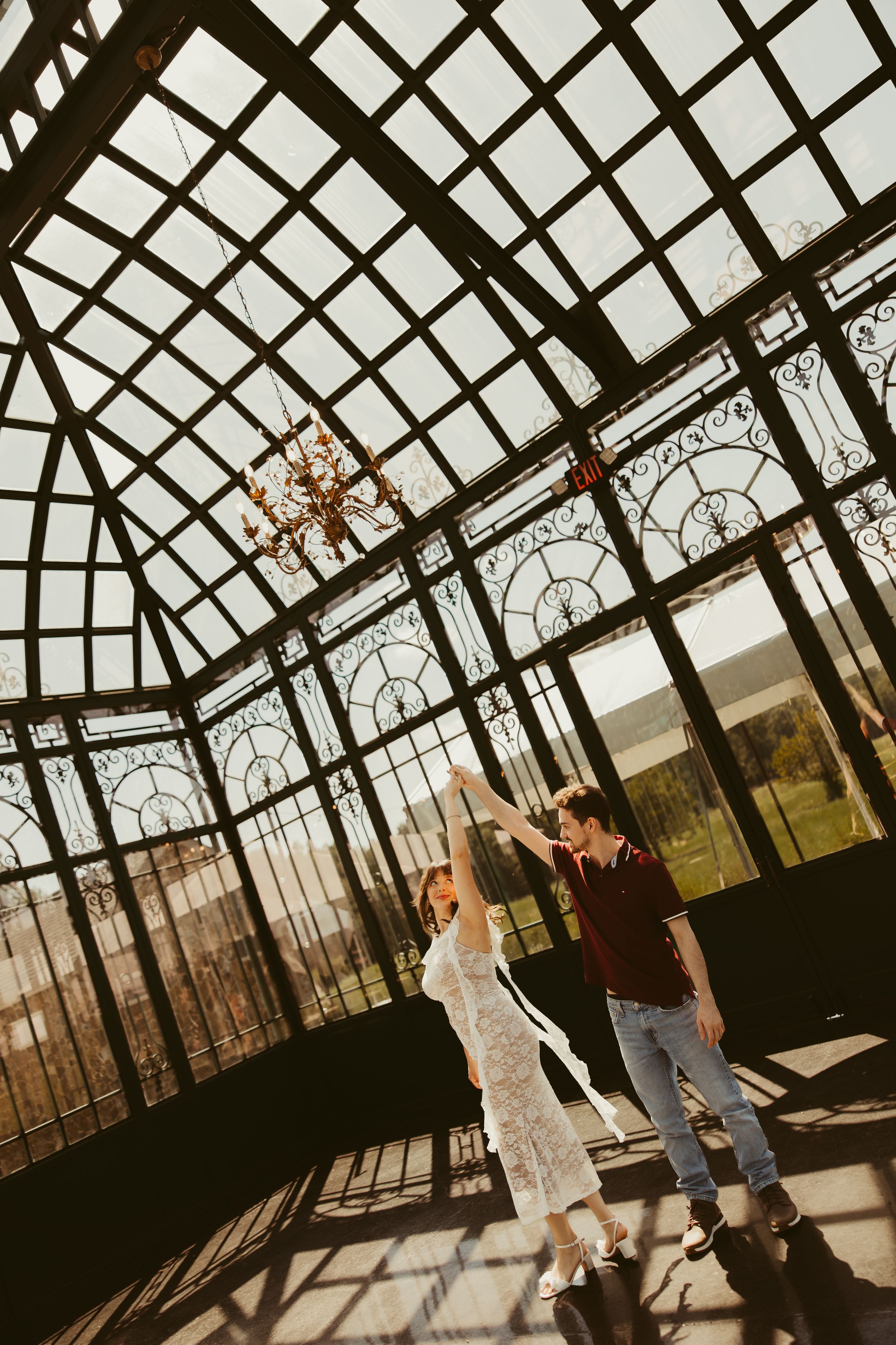 Couple dancing in a sunlit glass conservatory with ornate ironwork ceiling and chandelier.