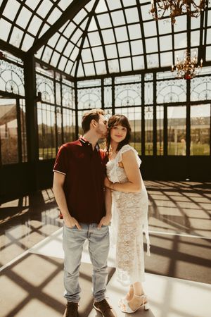 Couple posing in a sunlit glass greenhouse, man kissing woman's cheek, she wears a white lace dress.