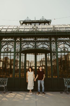 Couple holding hands standing in front of an ornate Victorian iron and glass greenhouse structure.
