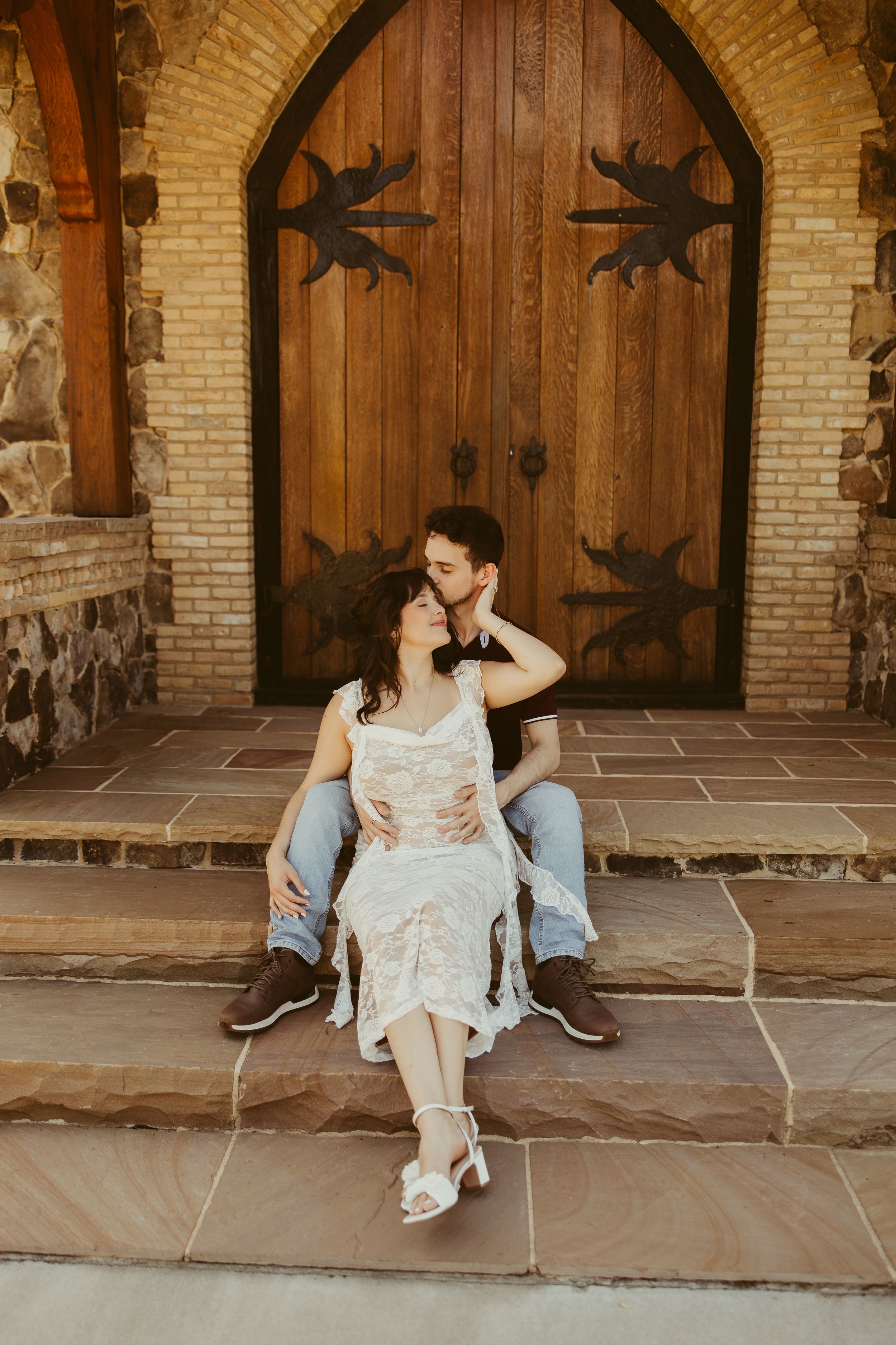 Couple sitting on stone steps before ornate wooden church doors, sharing a romantic moment in wedding attire.