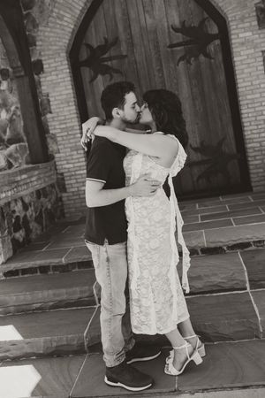 Black and white photo of a couple embracing and kissing in front of a rustic wooden door on stone steps.