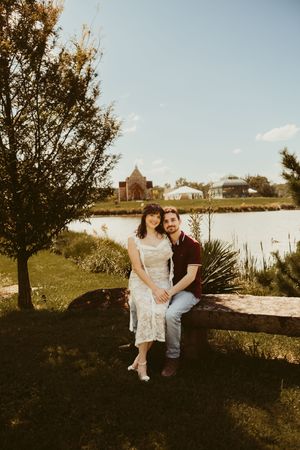 Couple sitting on a wooden bench by a serene lake, surrounded by lush greenery on a sunny day.