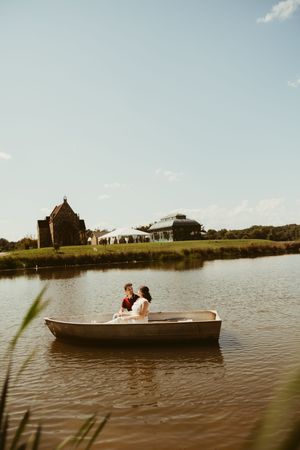 Couple in wedding attire sitting in a small rowboat on a calm lake with a historic chapel and greenhouse venue behind them.
