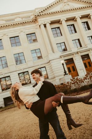 Man lifting woman in his arms laughing in front of a grand neoclassical building during an engagement photo session.