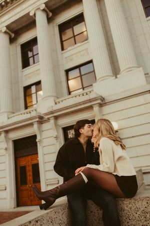 Couple sharing a romantic kiss while sitting on steps of a neoclassical building with tall columns at dusk.