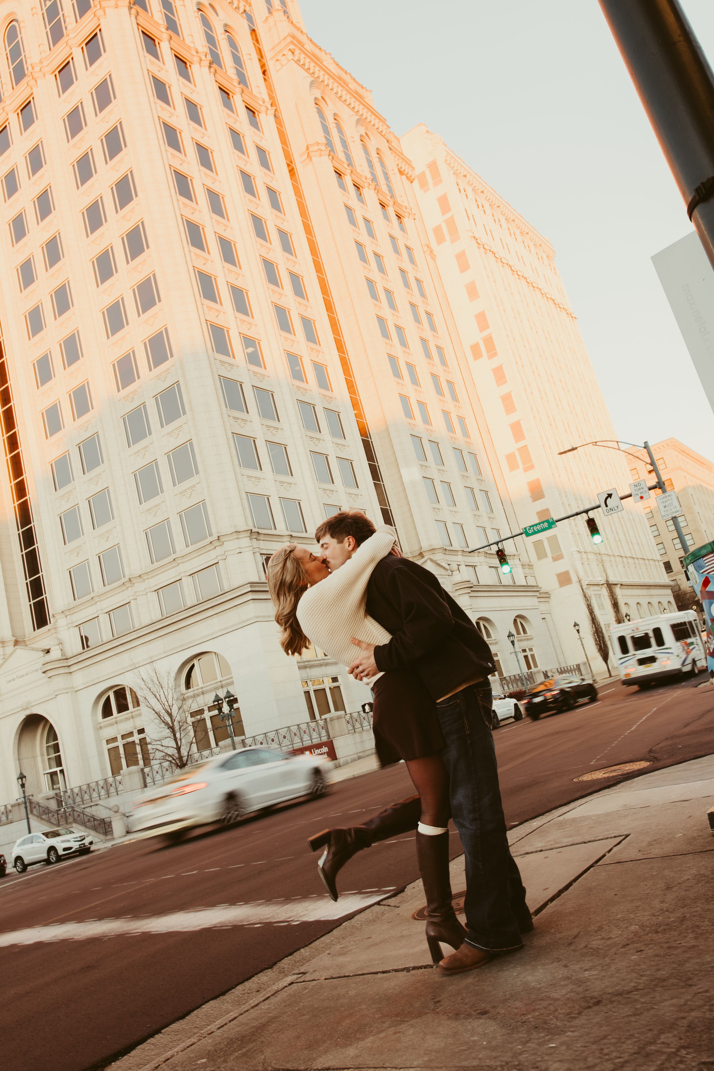 Couple sharing a romantic dip kiss on a city street corner with tall buildings glowing in golden sunset light.