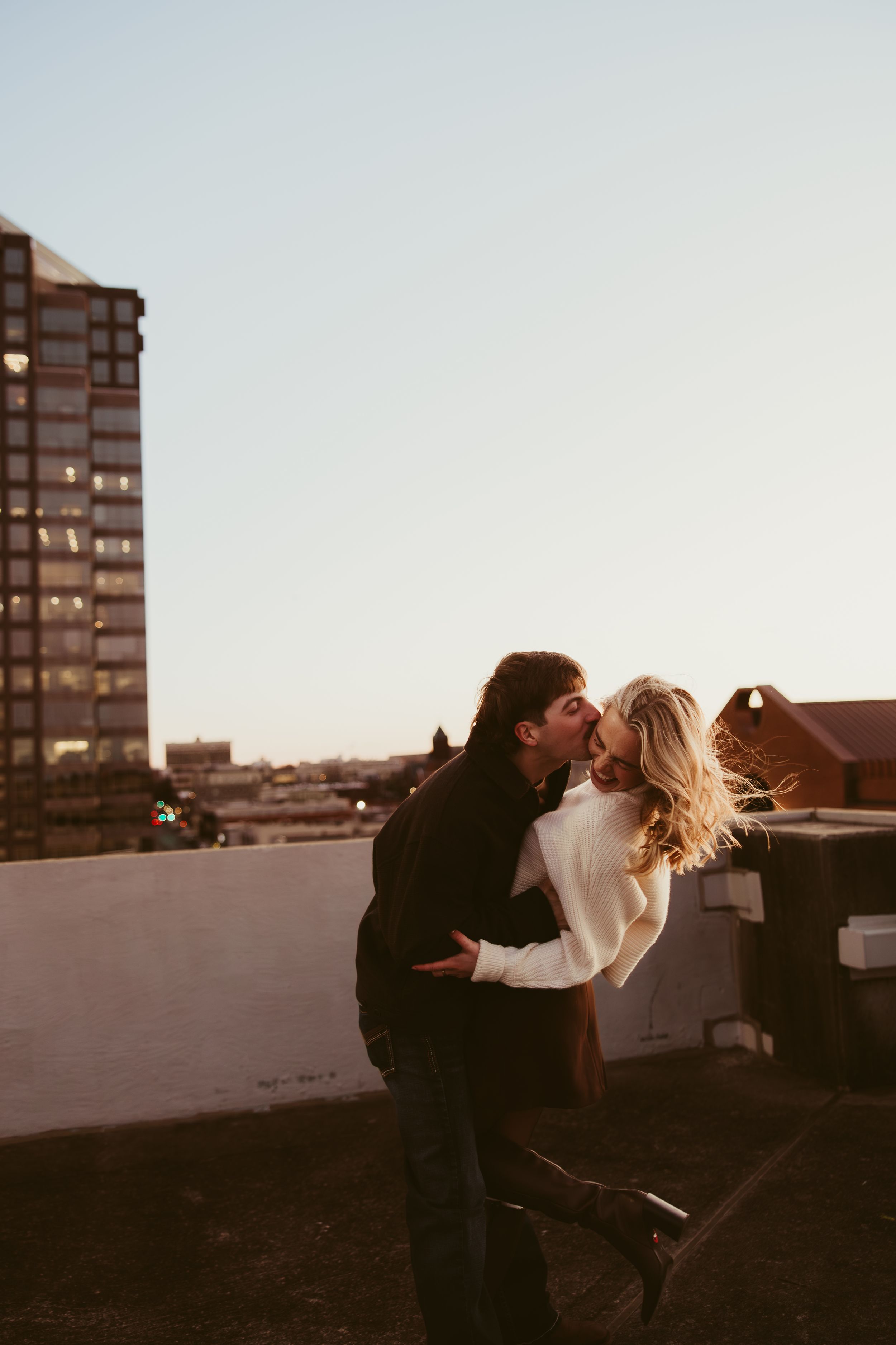 Couple sharing a passionate kiss on a rooftop at sunset with a city skyline in the background.