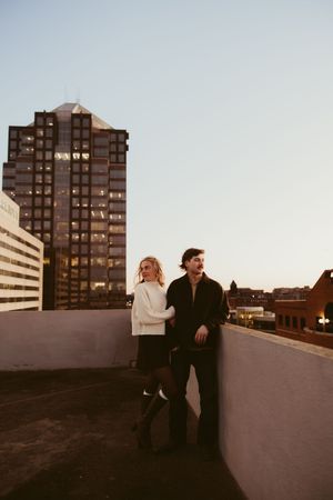 Couple standing together on a rooftop at dusk with a glowing city skyscraper behind them.