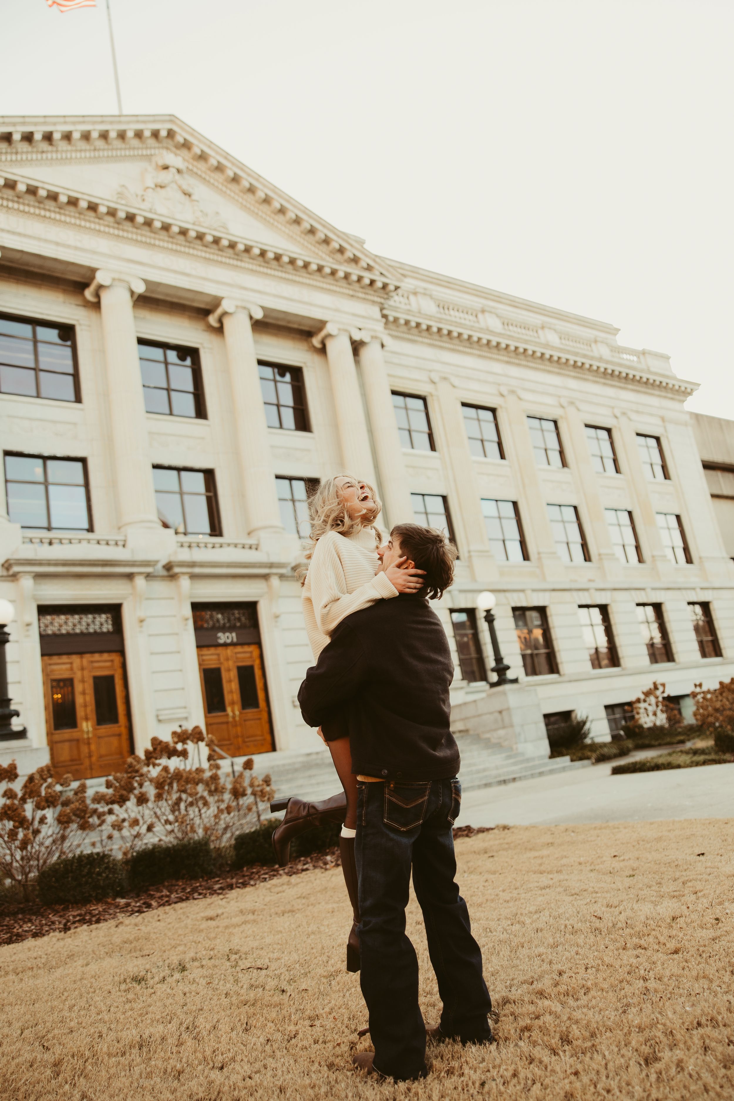 Man joyfully lifting laughing woman in front of a grand neoclassical building.