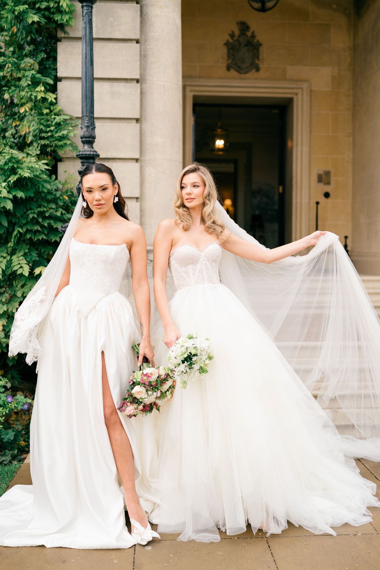 Two brides in elegant white wedding gowns pose together with flowing veils on a grand estate porch.