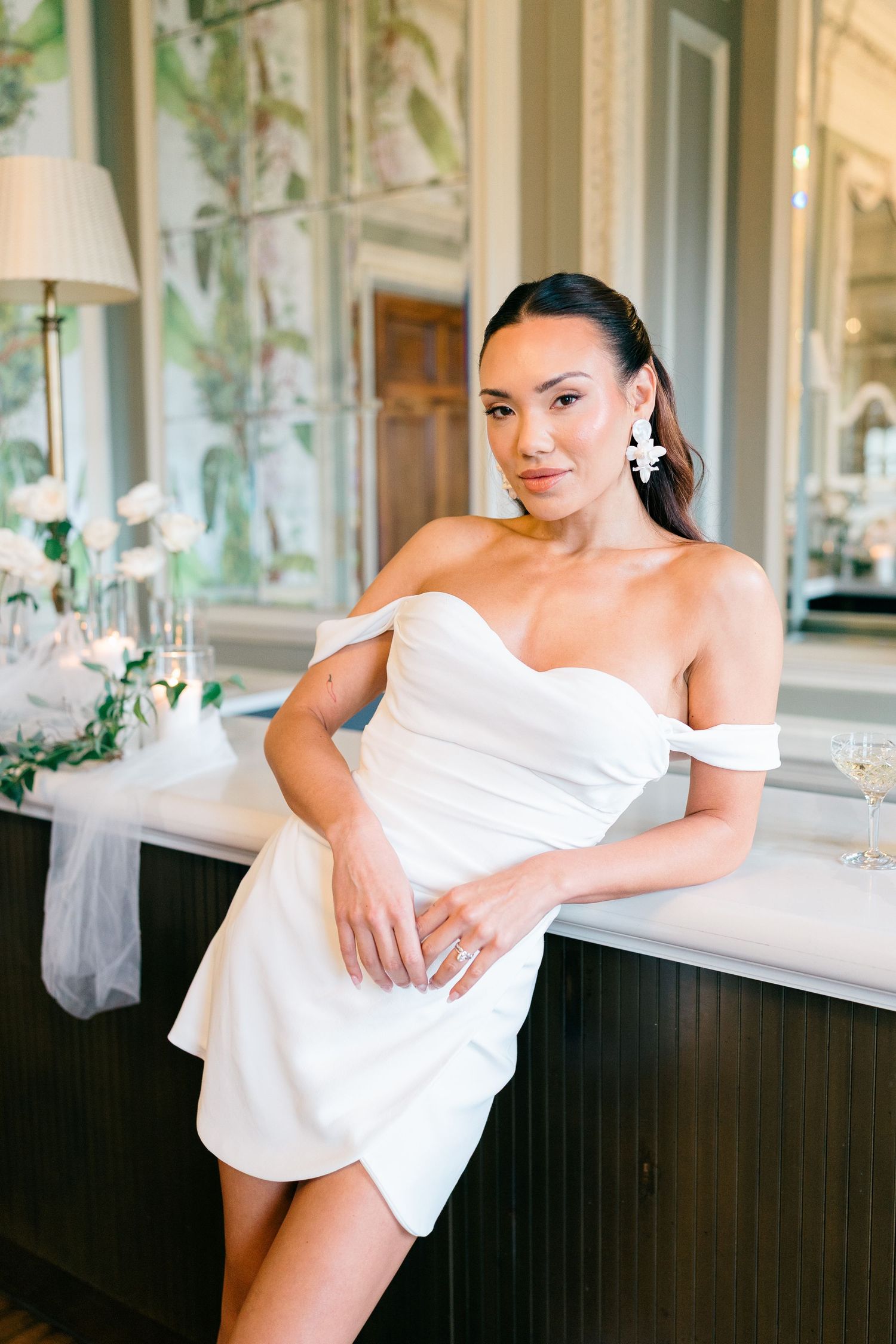 Elegant off-shoulder white mini dress worn in a luxurious room with ornate wallpaper and classic decor.