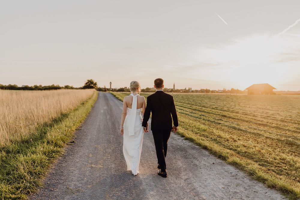 Paarshooting am Hochzeitstag bei Sonnenuntergang - das Brautpaar läuft Hand in Hand einen Feldweg entlang.