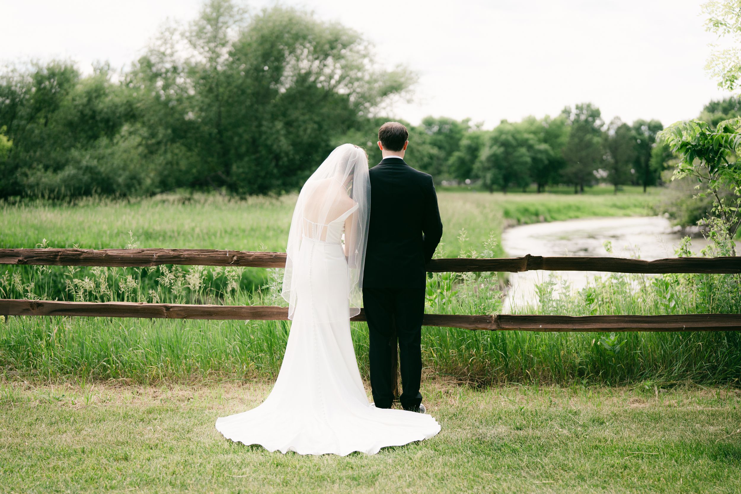 Bride and groom stand hand-in-hand facing a rustic wooden fence overlooking a lush green meadow on their wedding day.