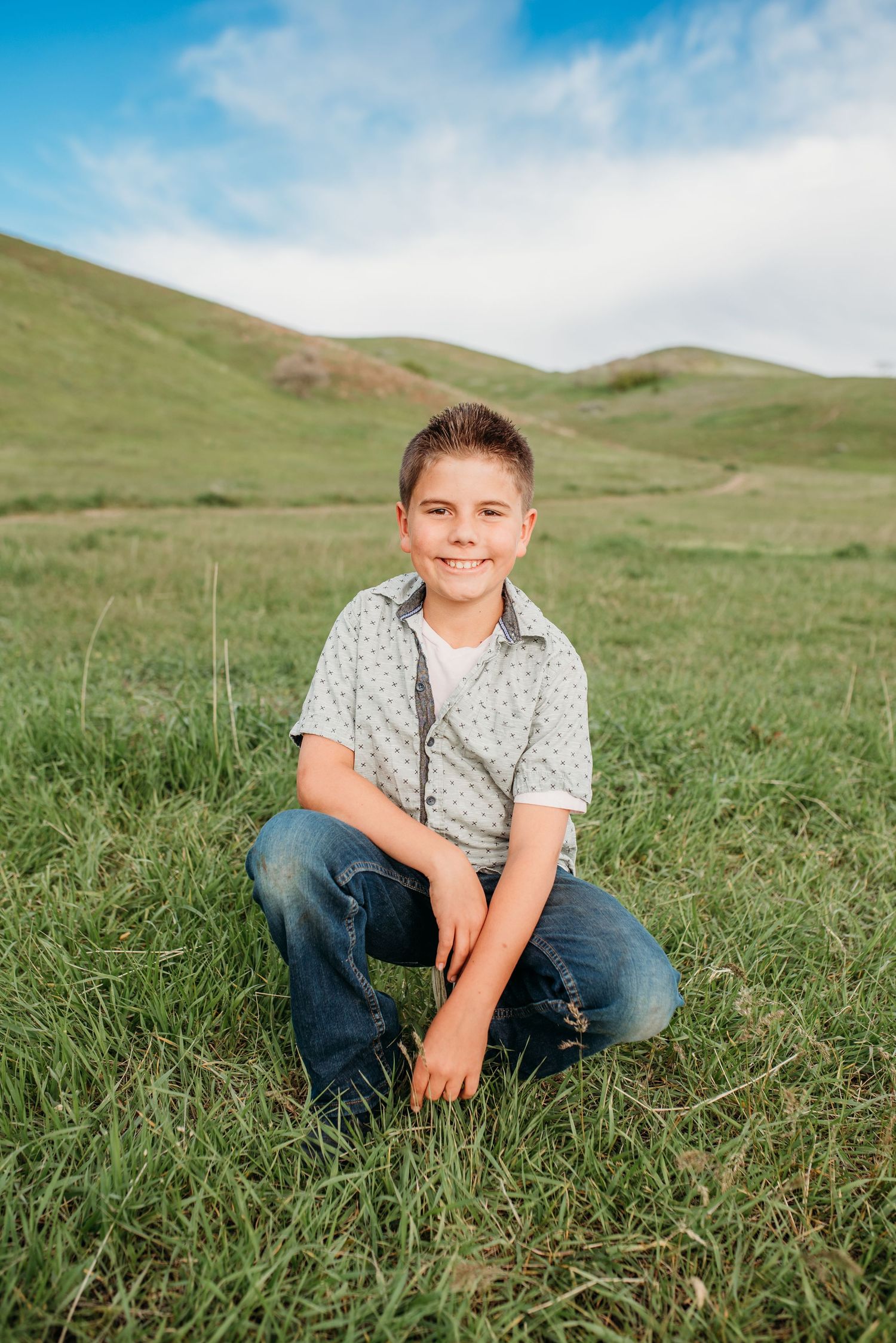 A child sits in green grass with rolling hills and blue sky in the background.