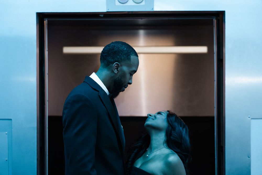 Two people in formal attire stand in a dimly lit elevator with blue lighting and reflective surfaces.