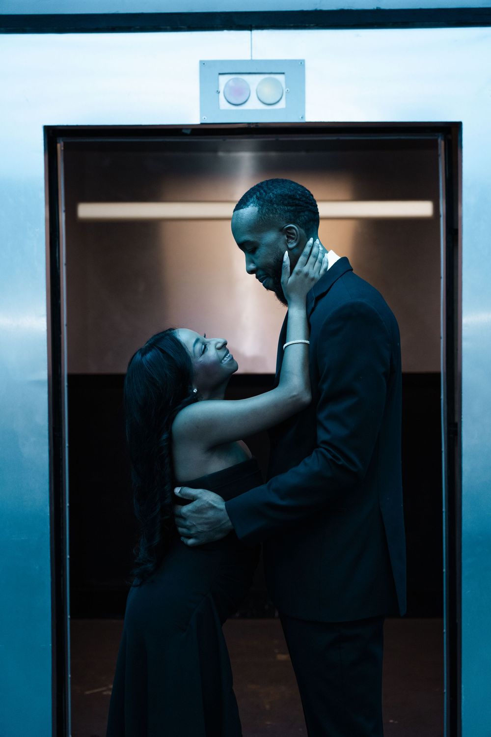 A romantic couple embraces intimately in a dimly lit elevator with blue tinted lighting and dramatic shadows.