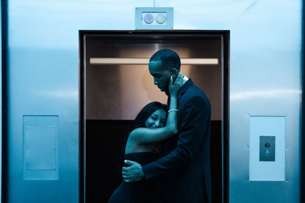 Two people embrace in a dimly lit elevator with blue-tinted lighting creating a moody atmosphere.