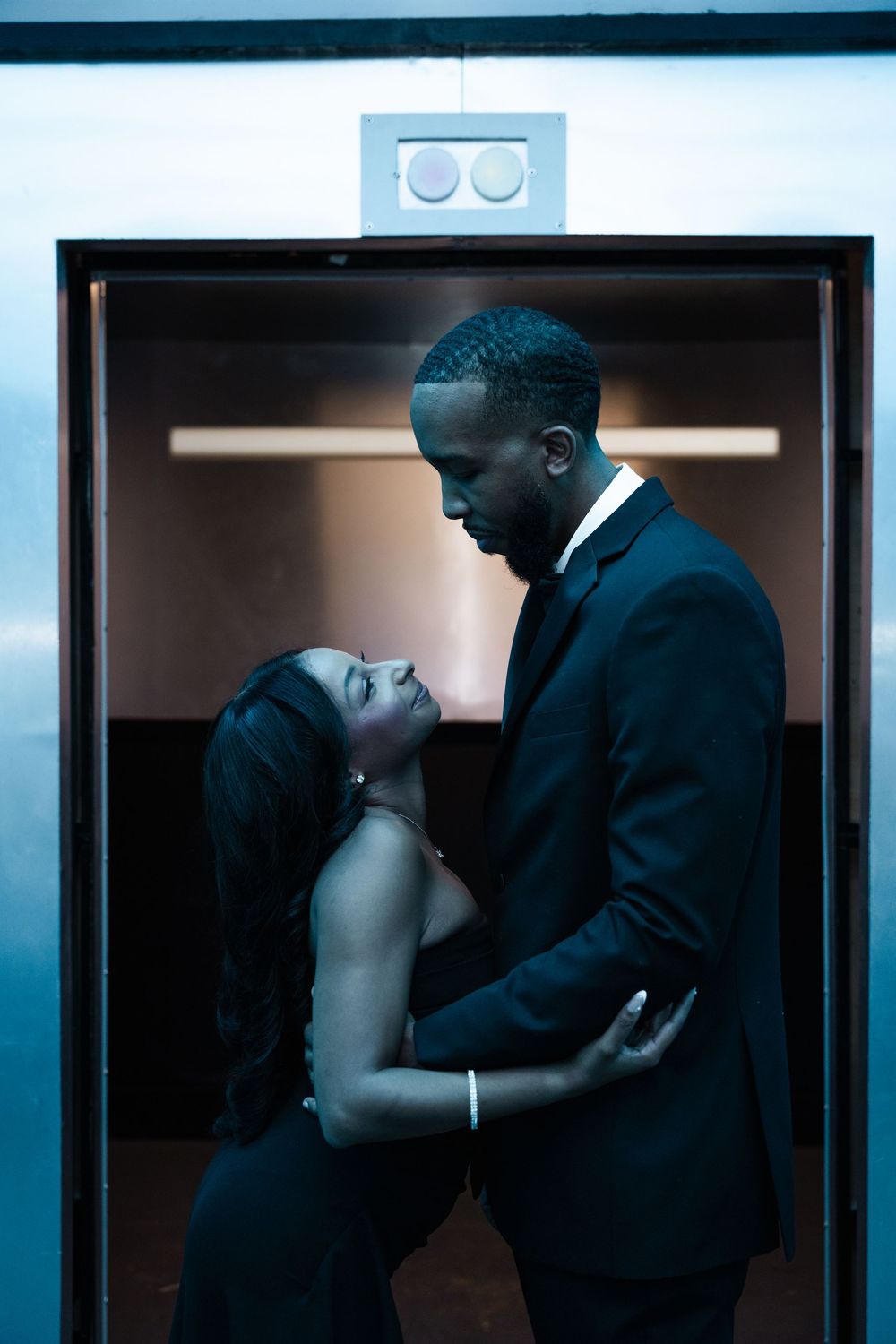 Romantic couple in formal attire share an intimate moment in a dimly lit elevator with blue tones.