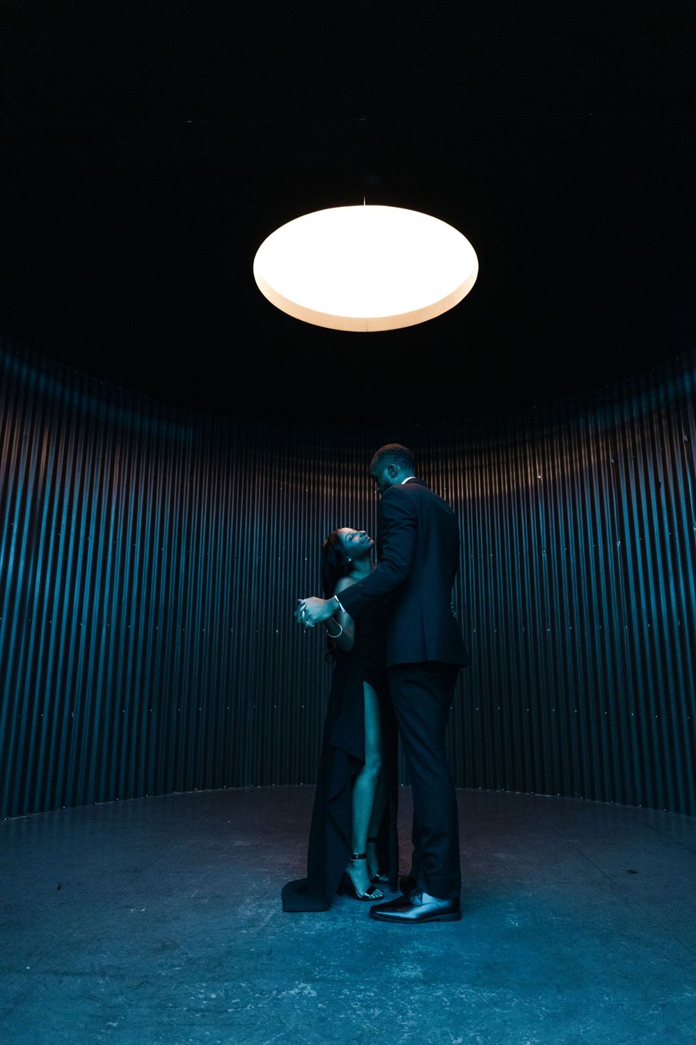 Two figures in dark coats stand beneath a bright circular light in a curved industrial space with blue tones.