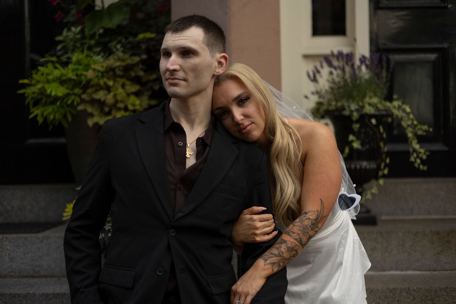 A couple in formal attire embrace outside a building, with the woman in white leaning on her partner in black.