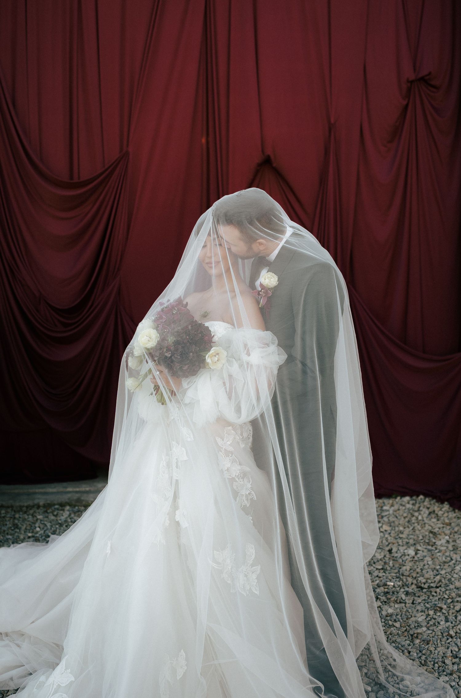 Ethereal white wedding dress with flowing veil photographed against deep burgundy drapes and scattered flower petals.