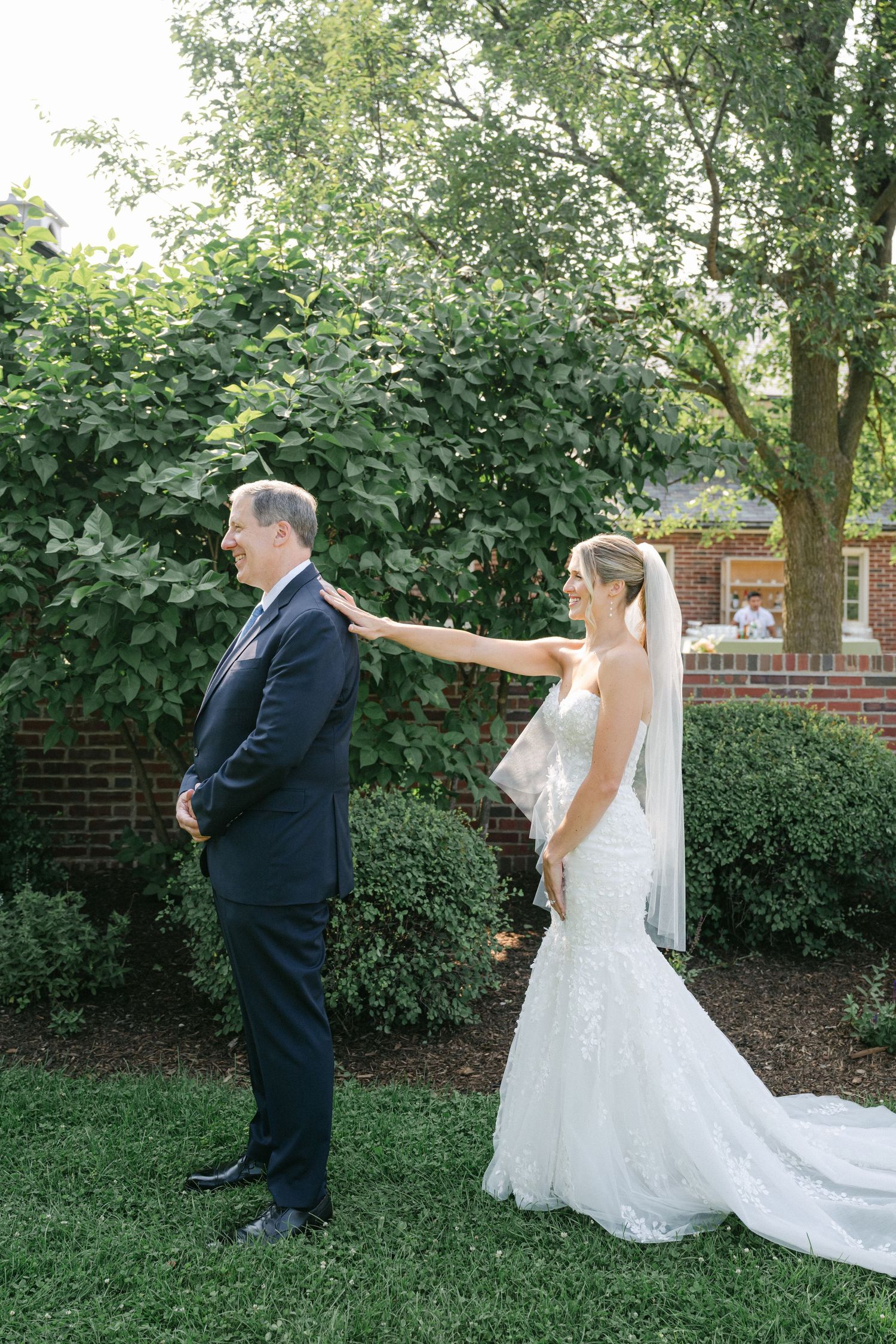 A couple in formal wedding attire share a playful moment in a garden setting with brick walls and greenery. - Elawa Farm