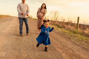 A child in a blue dress dances on a country road while parents watch during sunset.