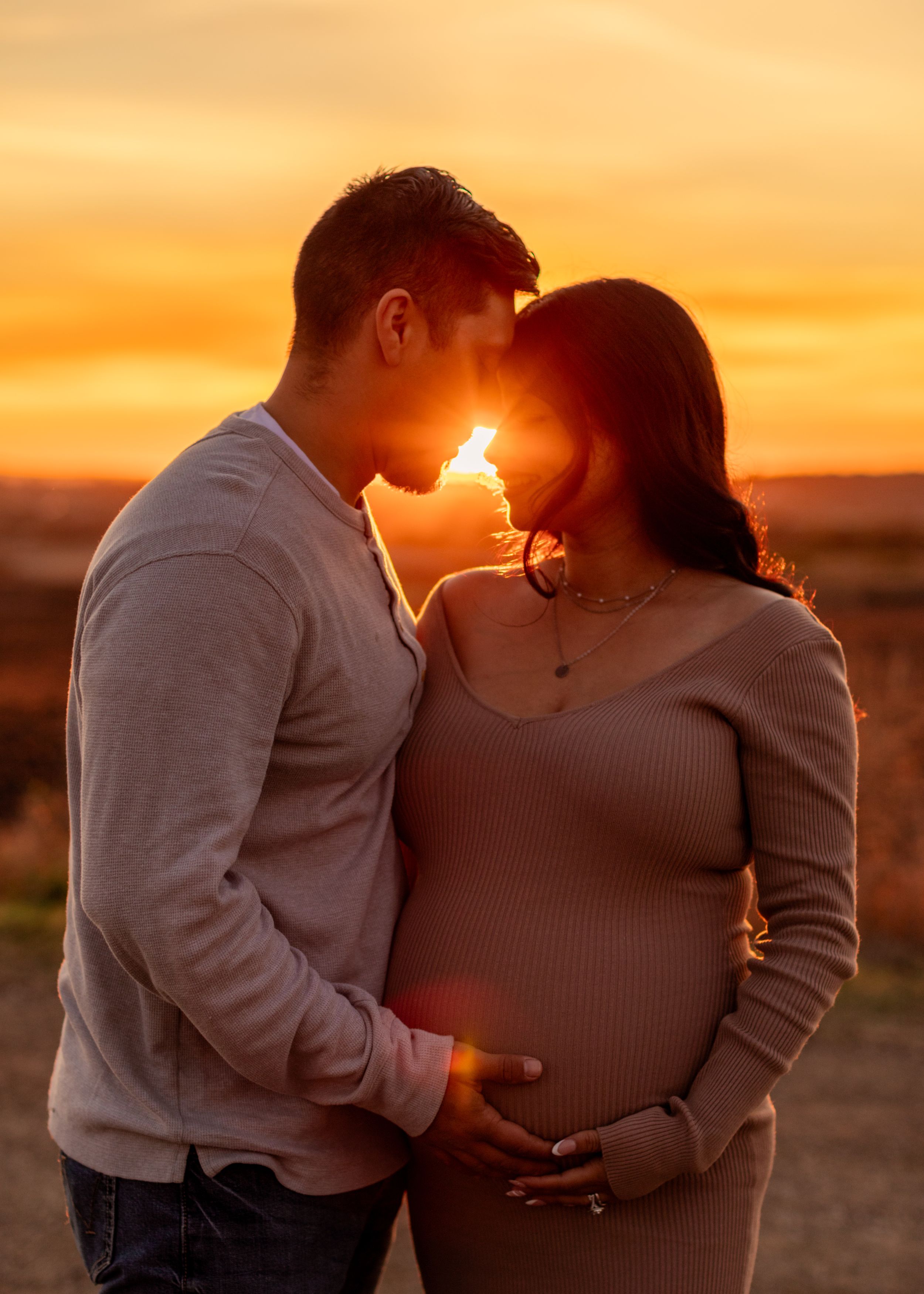Silhouetted couple shares intimate moment at sunset as they embrace and celebrate pregnancy in golden light.