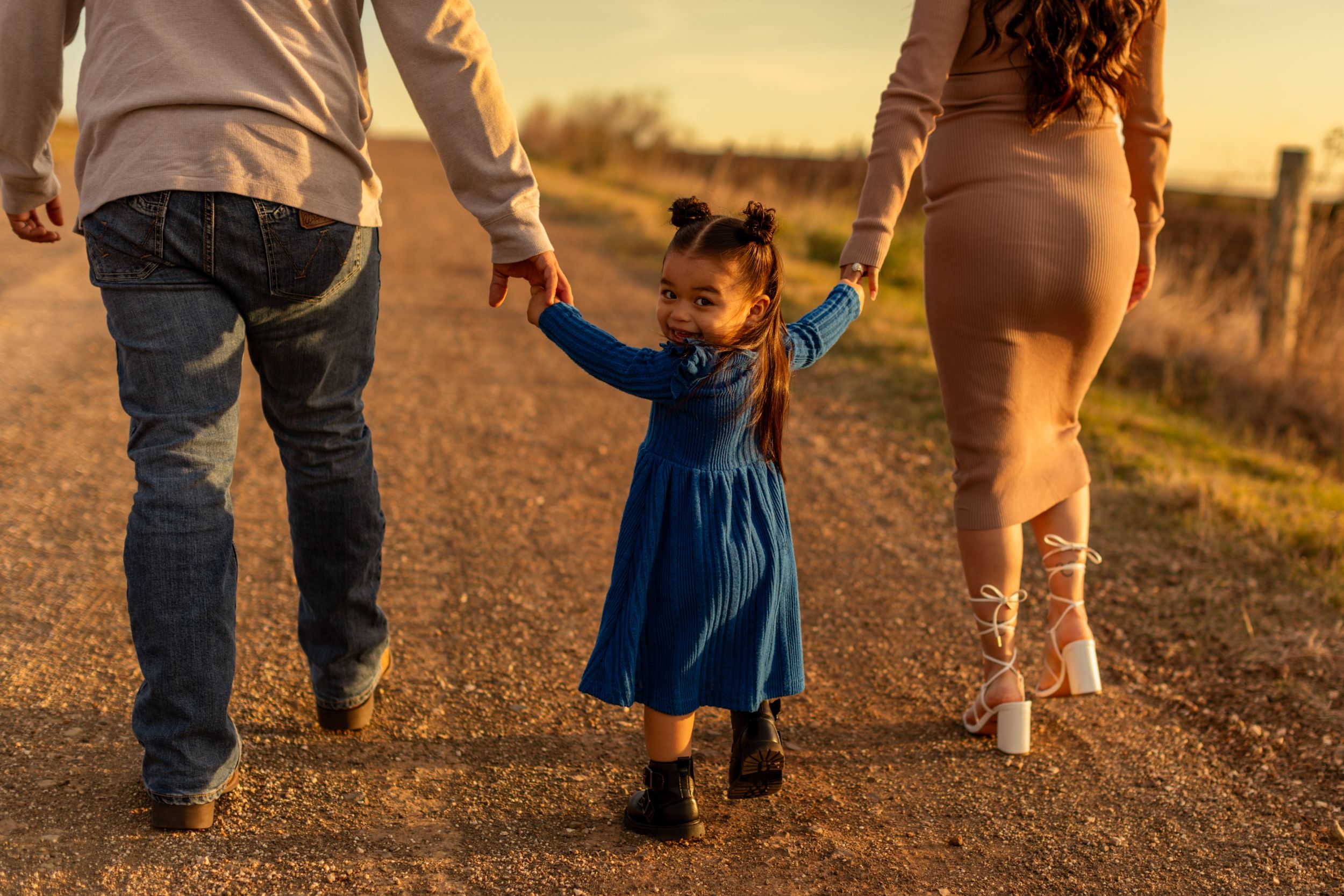A small child in a blue dress walks between two adults on a country road at sunset.