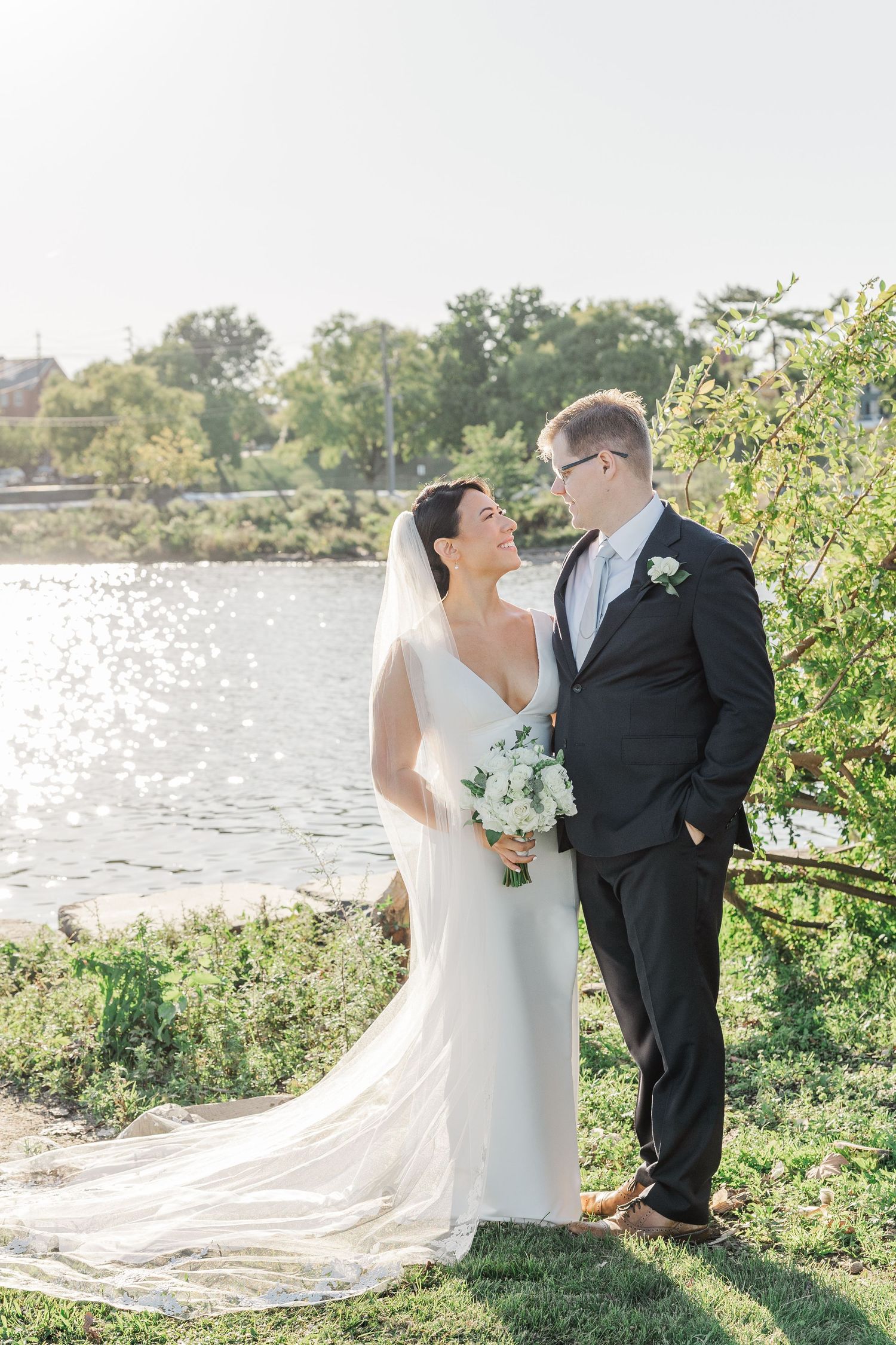 Newlywed couple shares romantic moments by a lakeside garden with flowing white wedding dress and classic black tuxedo.