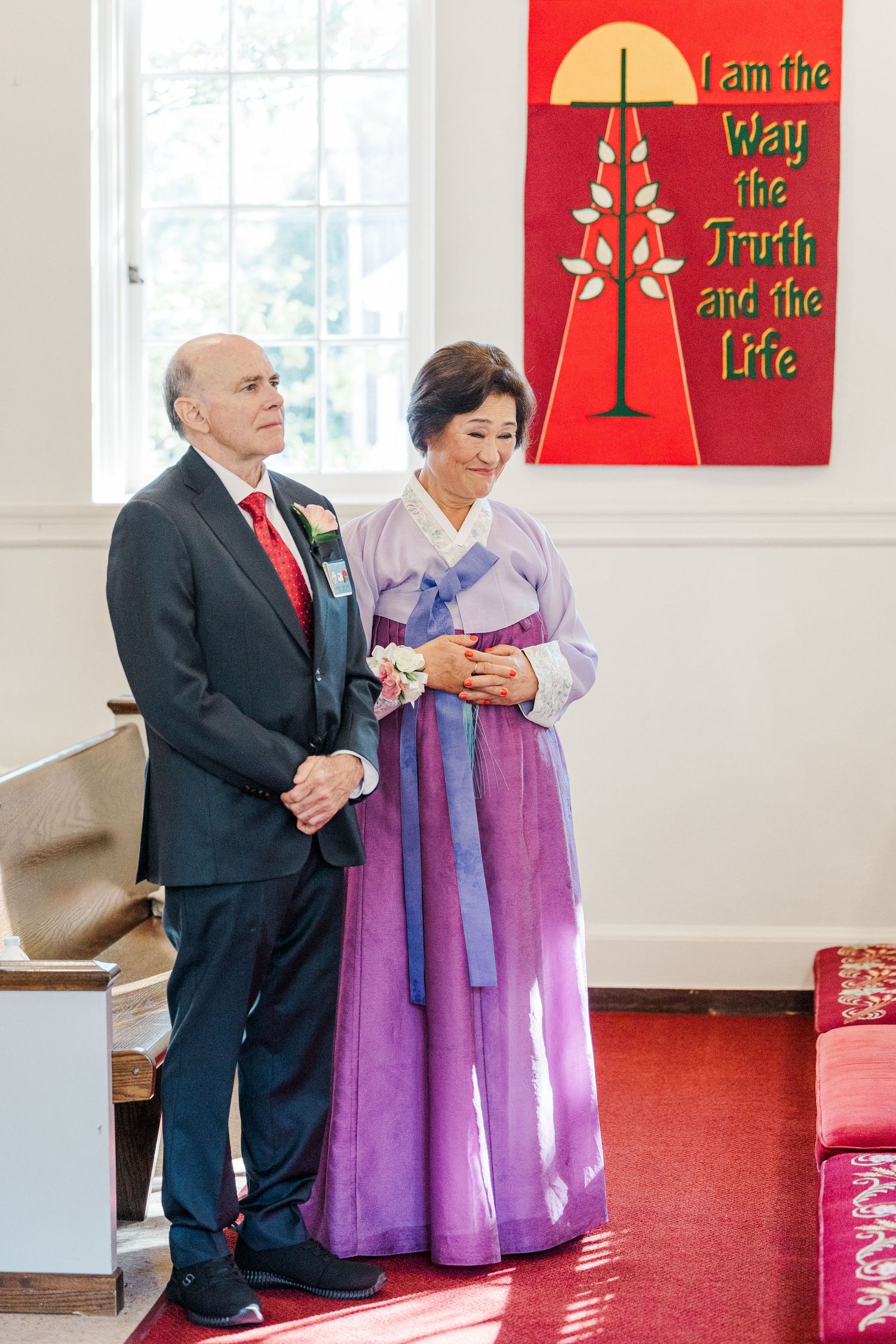 A couple in traditional Korean hanbok and western formal attire stand together in a church setting with red carpeting.