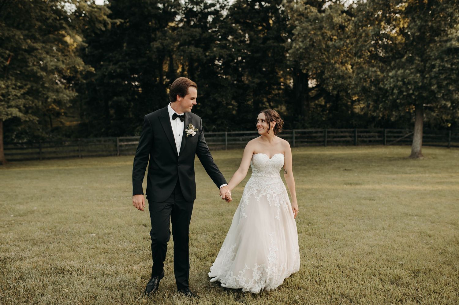 Bride and groom walk hand in hand across lush green field, she in lace gown, he in black tuxedo.