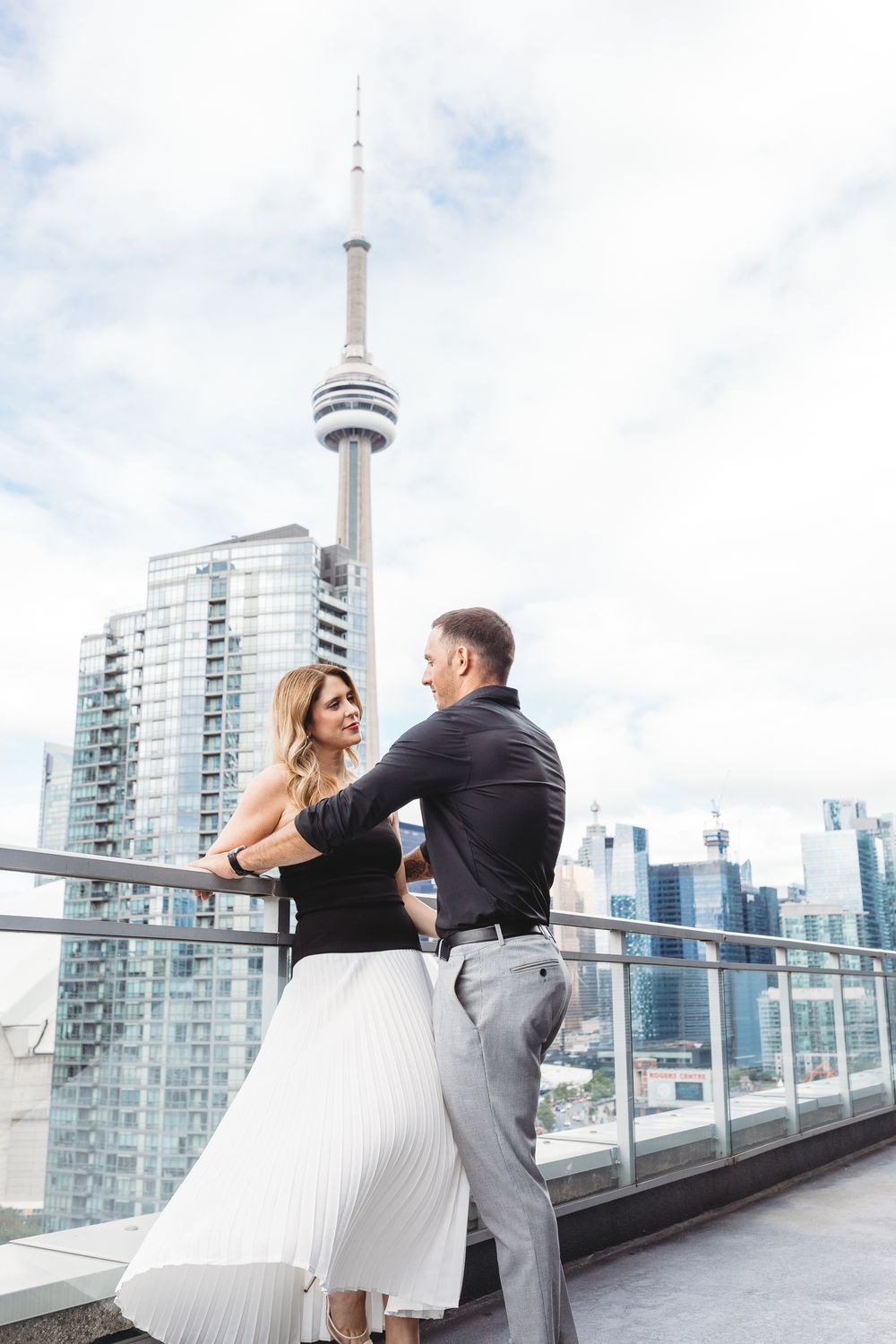 Couple performing dance steps on rooftop patio with glass railings and Toronto skyscrapers in view.