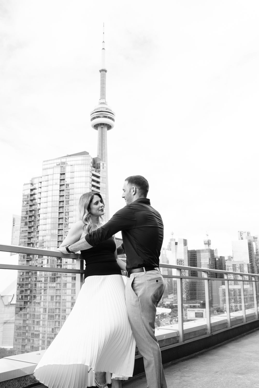 Black and white photo of couple embracing on rooftop terrace with city skyline backdrop.